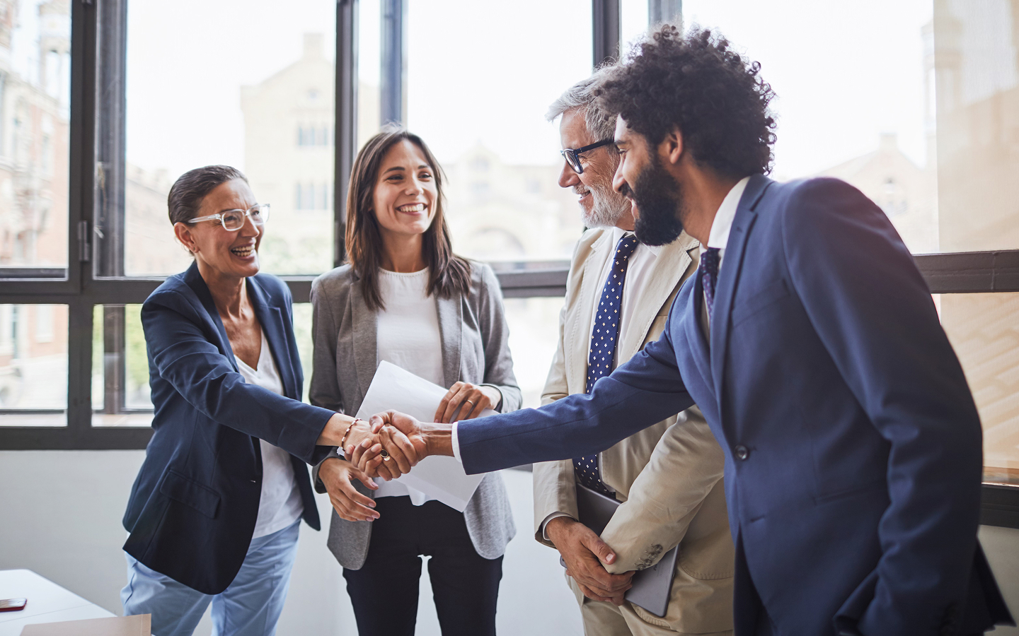Four people in business attire standing near window, two shaking hands while others observe.